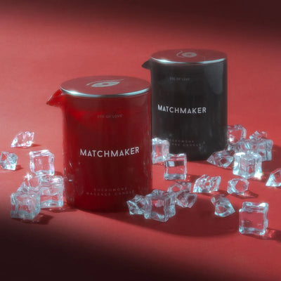 Two Eye of Love Matchmaker massage candles in red and black containers surrounded by decorative clear ice cubes on a red surface.