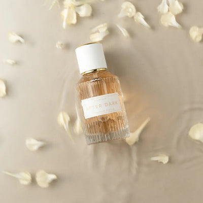 Elegant Eye of Love After Dark pheromone parfum bottle surrounded by soft white flower petals on a neutral background.
