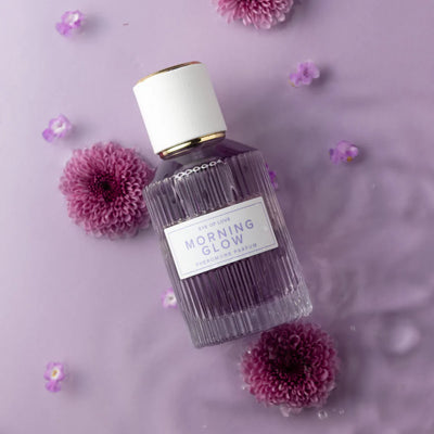 Eye of Love Morning Glow pheromone perfume bottle on lavender background with purple flowers and water droplets.
