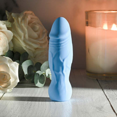 Blue silicone realistic vibrator with textured veins, standing upright beside white roses and a lit candle on a wooden surface.