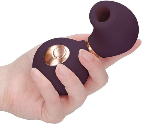 Hand holding a compact, dark purple clitoral vibrator with a rose gold button and a rounded, hollow silicone tip.