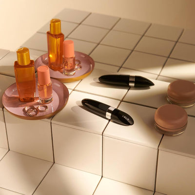 Two black Lelo Mia 2 massagers displayed on white tiled steps alongside cosmetic bottles, rings, and small containers.