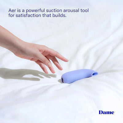 Hand reaching for a compact lavender Dame Aer Air Pulse Stimulator on a white bedspread with product description text.