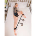 Person lying on bed wearing black bodysuit with Saffron Spreader Bar and red-black adjustable cuffs on ankles and wrists.