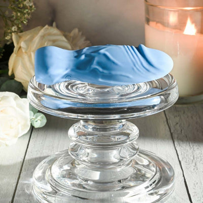 Blue silicone realistic vibrator displayed on a clear glass pedestal with white roses and a lit candle in the background.