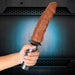 Hand holding a mocha-colored realistic dildo attached to a black Karbonite Lock On handle against a dark background.