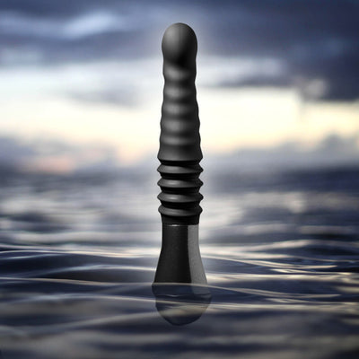 Black Temptasia Lazarus thrusting dildo with textured shaft standing upright in water against a blurred sky background.
