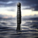 Black Temptasia Lazarus thrusting dildo with textured shaft standing upright in water against a blurred sky background.