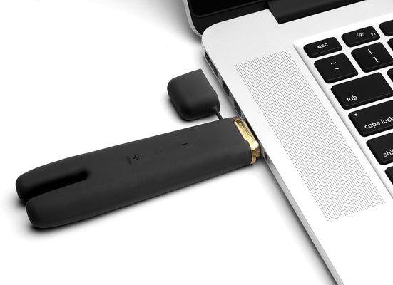 Black Crave Duet Flex dual-tip vibrator plugged into a laptop USB port for charging with attached protective cap.