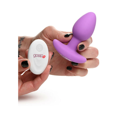 Hand holding a vibrant violet silicone butt plug and a white remote control with Gossip logo on a white background.