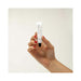 Hand with red nail polish holding a small white tube of Bijoux Indiscrets Slow Sex Oral Sex Balm against a plain background.