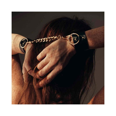 Close-up of hands behind head wearing black vegan leather wide cuffs connected by a gold chain from Bijoux Indiscrets Maze Collection.