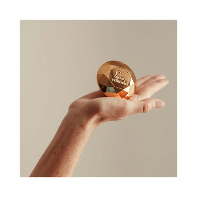 Hand holding a rose gold faceted diamond-shaped clitoral vibrator by Bijoux Indiscrets against a neutral background.