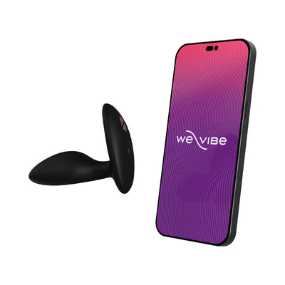 Black silicone vibrating anal plug next to smartphone displaying We-Vibe app interface for remote control.