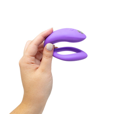 Hand holding a purple We-Vibe Sync O couples vibrator with flexible O-shaped design and control buttons.