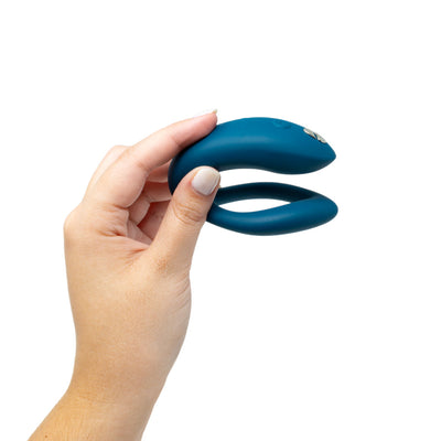 Hand holding a blue We-Vibe Sync O couples vibrator with a flexible O-shaped design against a white background.