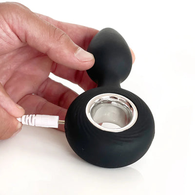 Hand holding a black silicone rechargeable anal plug with a silver ring, shown being plugged in for charging.