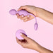 Hands holding a lilac vibrating kegel ball and matching wireless remote control against a pink background.