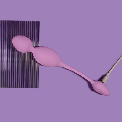 Lilac Wellness Raine vibrating kegel ball with smooth silicone surface on purple background, shown with charging cable attached.