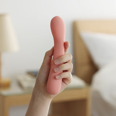 Hand holding a sleek, pink iroha mai TOKI personal massager with a smooth, curved design in a softly lit bedroom setting.