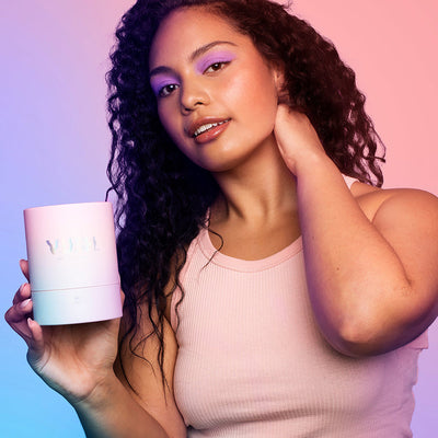 Woman with curly hair and purple eyeshadow holding a pastel pink Vush Gloss Bullet Vibrator packaging against a pink and blue background