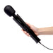 Hand holding a sleek black Le Wand Plugin vibrating massager with control buttons and attached power cord against a white background.