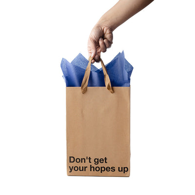 Hand holding a kraft gift bag with brown ribbon handles and blue tissue paper, printed with "Don't get your hopes up" text.