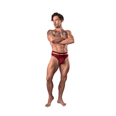 Muscular man wearing burgundy lace cutout strappy thong standing with arms crossed on white background.