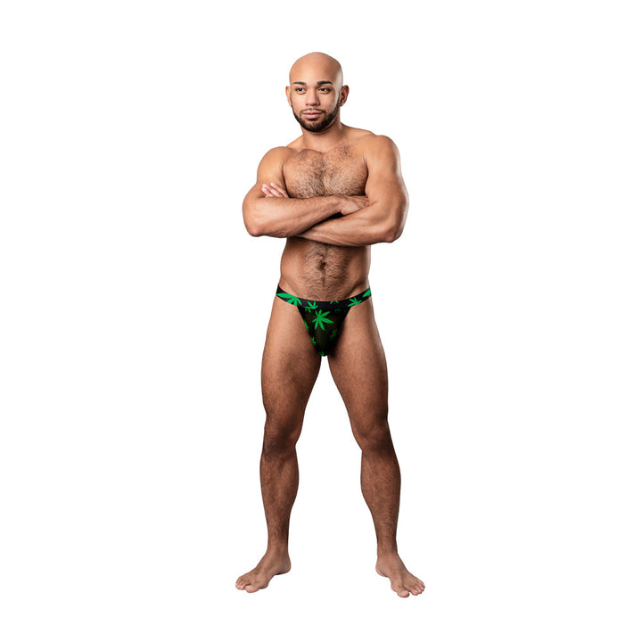 Male model wearing Male Power Hazy Dayz micro thong with green pot leaf print on black background, standing with arms crossed.