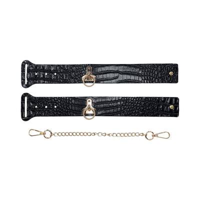 Black crocodile-textured leg cuffs with gold-tone O-ring hardware and detachable gold chain connector on white background.