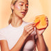Woman holding and demonstrating the orange VUSH Luna wearable vibrator with its compact charging case against a peach background.