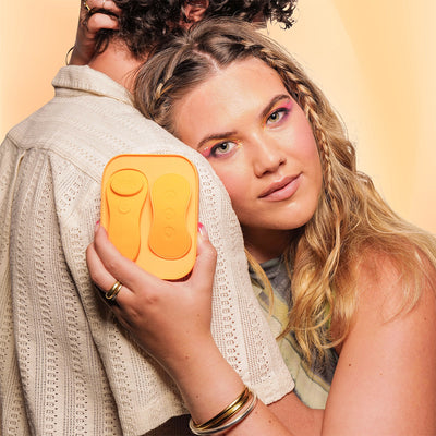 Woman with braided hair holding an orange VUSH Luna Wearable Vibrator remote and charging case while embracing a partner.