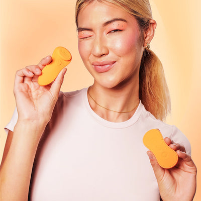 Smiling woman holding the bright orange VUSH Luna wearable vibrator and its matching remote against a peach background.