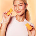 Smiling woman holding the bright orange VUSH Luna wearable vibrator and its matching remote against a peach background.