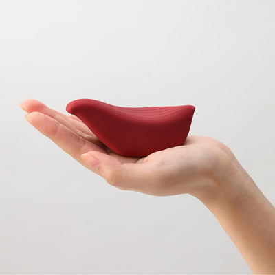 Hand holding a compact, bird-shaped iroha+ TORI Renewal vibrator in deep red silicone with smooth, curved design.