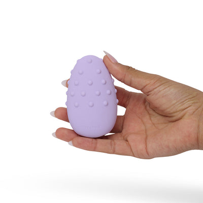 Hand holding a lilac Je Joue Mimi Deux double-sided clitoral vibrator with raised textured dots on one side.