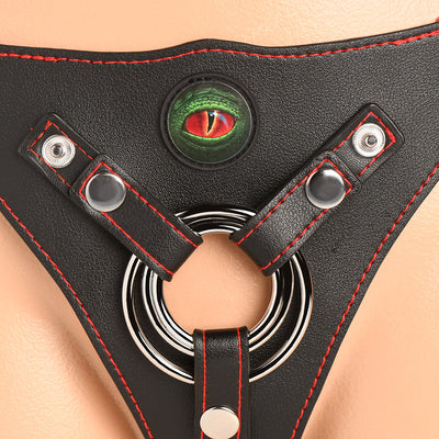 Close-up of black vegan leather strap-on harness with red stitching, metal O-rings, and a green creature eye embellishment.