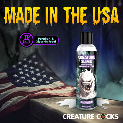 Bottle of Creature Slime unscented lubricant with monster-themed label, placed on wooden surface with spilled liquid and American flag background.