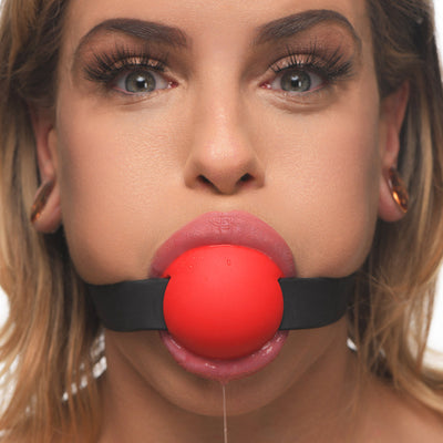 Close-up of a person wearing a red silicone ball gag with a black adjustable locking strap.