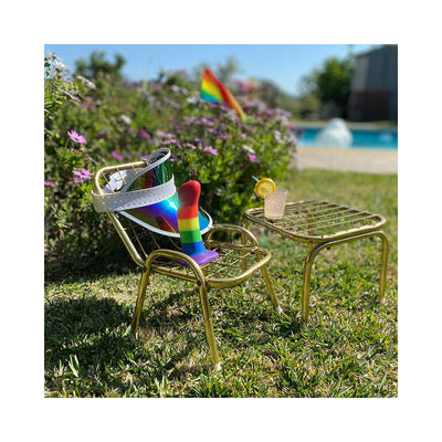 Rainbow silicone dildo displayed on a small gold chair outdoors with a matching table and a drink with lemon garnish.
