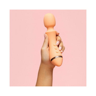 Hand holding a peach-colored VUSH Majesty 2 wand vibrator with control buttons against a pink background.
