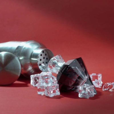 Black diamond-shaped Eye of Love Matchmaker perfume bottle with clear cap surrounded by ice cubes and a metal cocktail shaker on red background