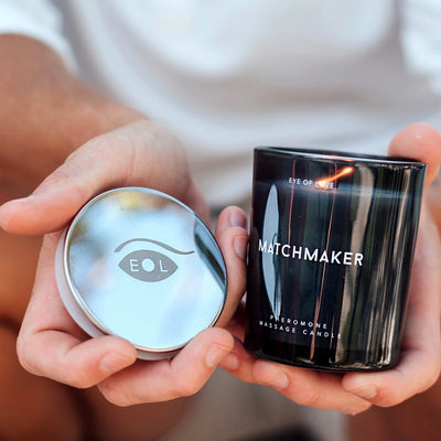Hands holding a black Eye of Love Matchmaker pheromone massage candle with a silver lid featuring the EOL logo.