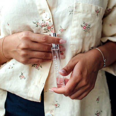 Hands holding a slim clear bottle of Eye of Love Unscented Pheromone Perfume against a floral patterned shirt background.