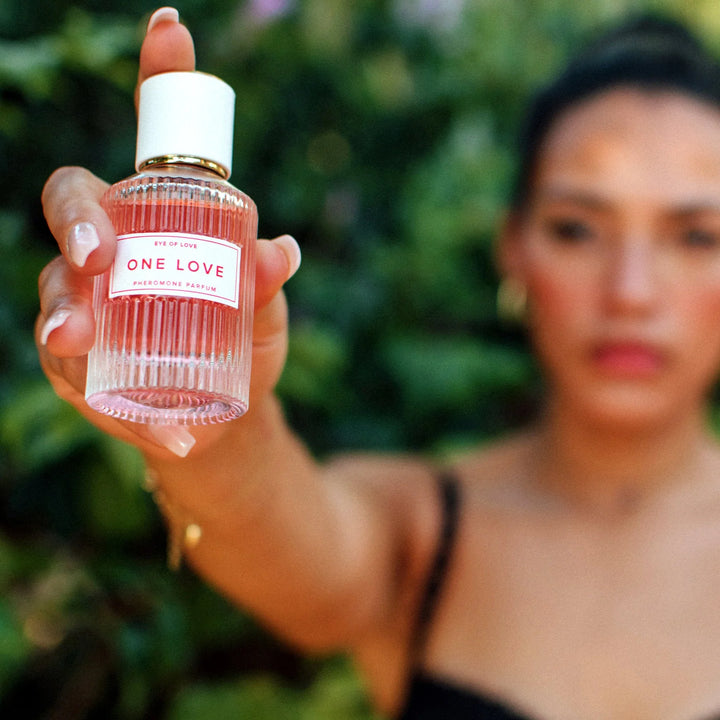 Hand holding Eye of Love One Love pheromone parfum bottle with blurred natural green background.