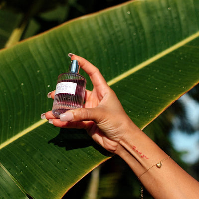 Hand holding Eye of Love Morning Glow pheromone perfume bottle against a large green leaf background.