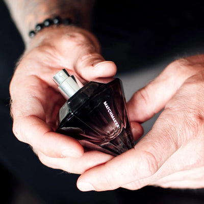 Hands holding a black diamond-shaped Matchmaker pheromone perfume bottle with a silver spray top.