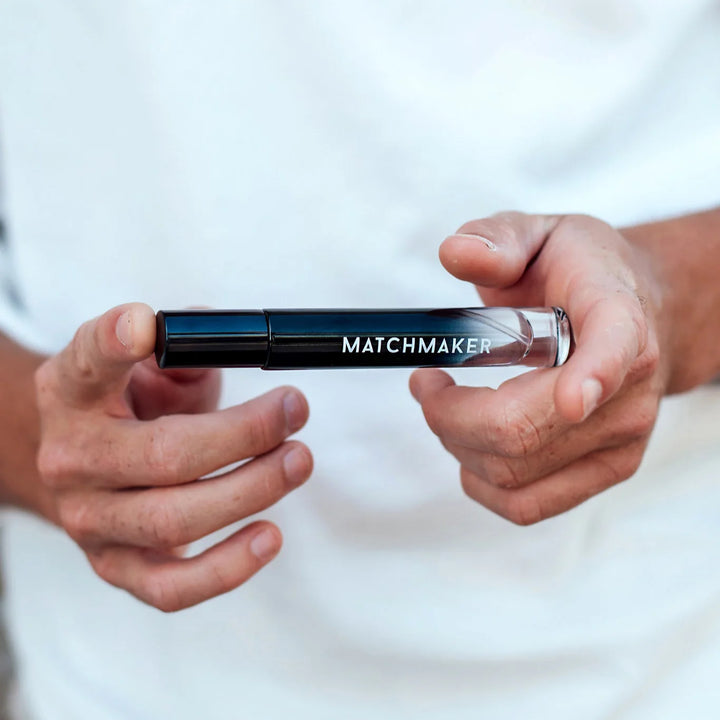 Hands holding a sleek black and clear 10ml Matchmaker pheromone perfume roller bottle against a white background.