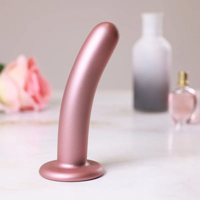 Rose gold smooth silicone G-spot dildo with suction base displayed on marble surface with blurred rose and bottles in background.