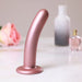 Rose gold smooth silicone G-spot dildo with suction base displayed on marble surface with blurred rose and bottles in background.